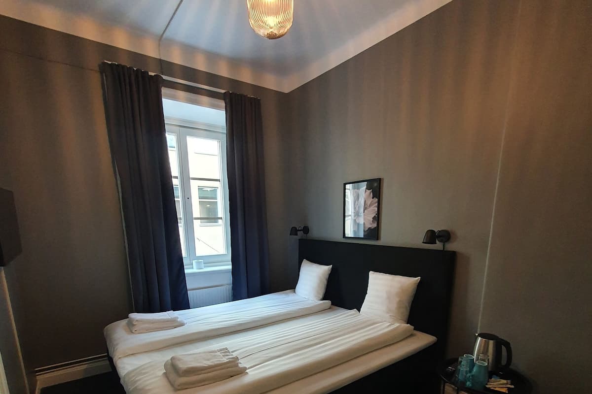 A hotel room with a large bed, a bedside table with a kettle, and a window with curtains.