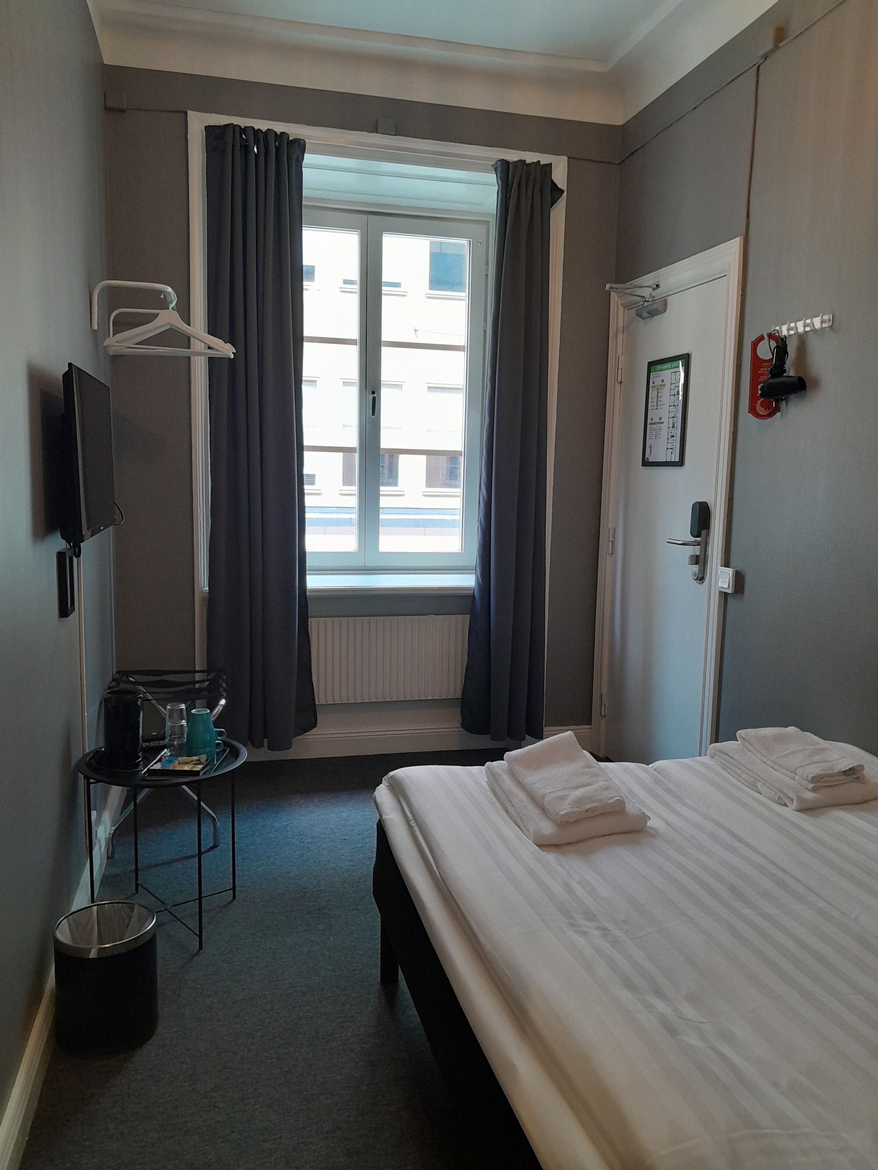 Una habitación de hotel con una cama, una mesita con tetera y tazas, un televisor, una ventana con cortinas, una puerta con un cartel y un radiador.