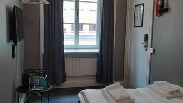 Una habitación de hotel con una cama, una mesita con tetera y tazas, un televisor, una ventana con cortinas, una puerta con un cartel y un radiador.