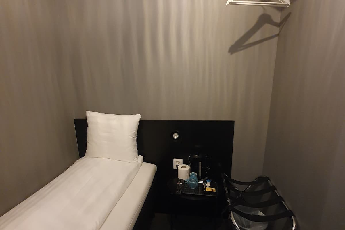A small hotel room with a single bed, a bedside table with a lamp, a chair, and a hanging coat rack.