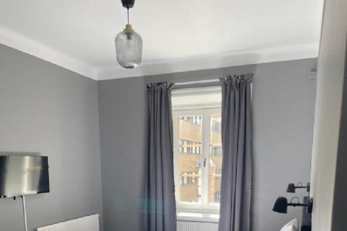 A hotel room with a bed, a bedside table, a lamp, a window with curtains, and a view of buildings outside.
