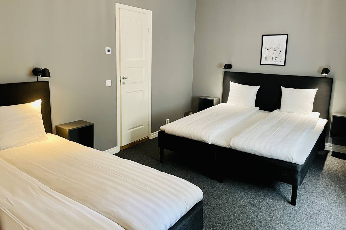 A hotel room with two beds, a door, a framed picture on the wall, and a nightstand with a lamp.