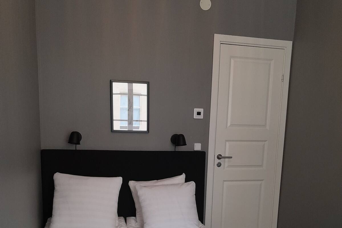 A neatly made bed with white linens and towels, a dark headboard, a small window, and a door to the right.