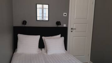 A neatly made bed with white linens and towels, a dark headboard, a small window, and a door to the right.