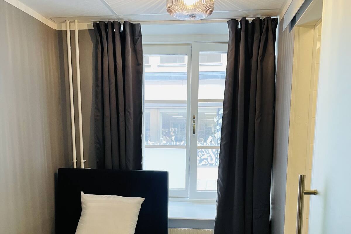 A small, neatly arranged bedroom with a single bed, a window with curtains, a radiator, and a door.
