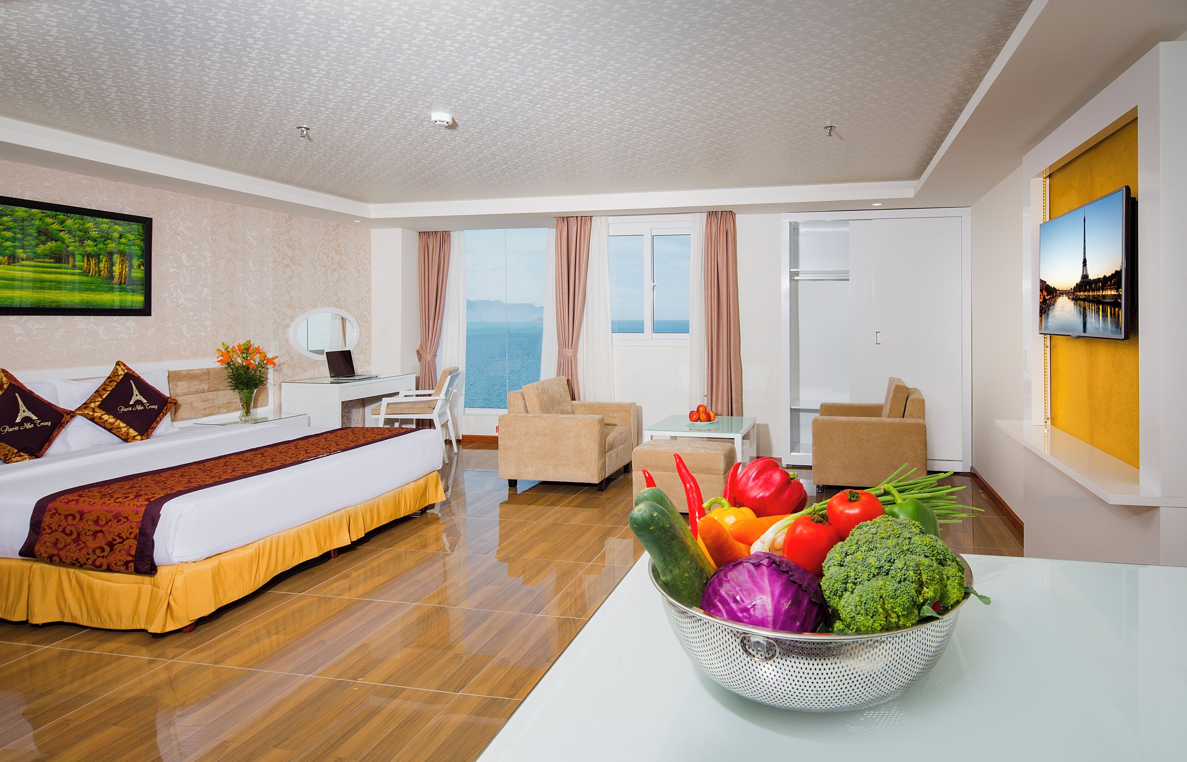 A hotel room with a bed, a dining table with a fruit bowl, a chair, a sofa, a TV, and a view of the sea.
