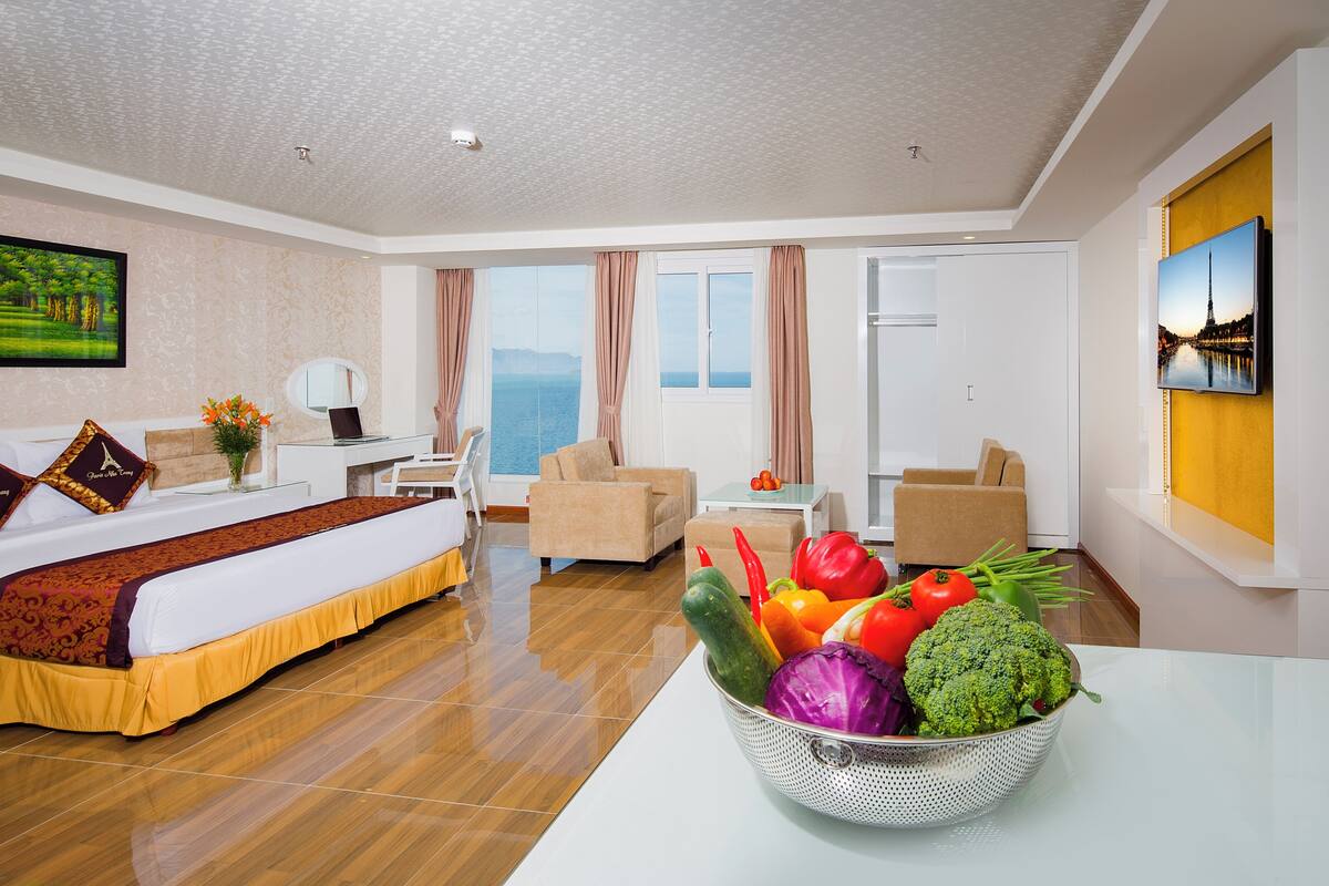 Une chambre d’hôtel avec un lit, une table à manger agrémentée d’un panier de fruits, une chaise, un canapé, une télévision et une vue sur la mer.