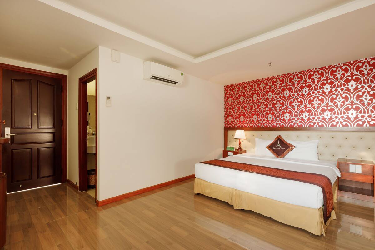A hotel room with a wooden floor, a large bed with a red and white bedspread, a headboard with a red patterned design, and a nightstand with a lamp.