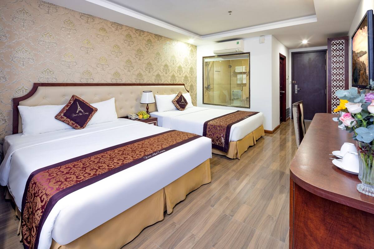 A hotel room with two beds, a wooden floor, a bathroom with a glass door, and a desk with a vase of flowers.