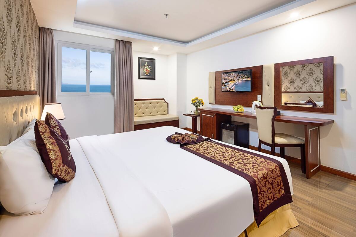 A hotel room with a large bed, a desk, a chair, a TV, and a view of the sea.