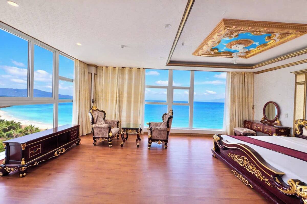 A room with a large window overlooking a beach, a bed, a dresser, and two armchairs.