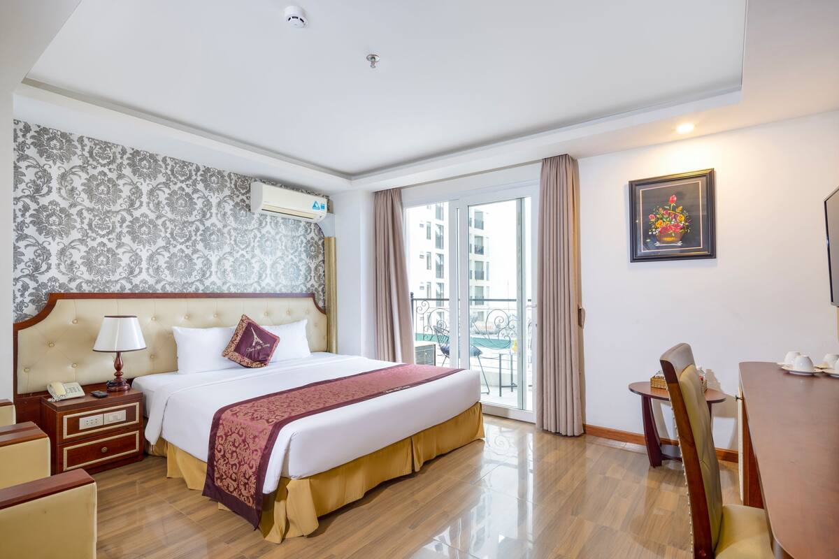 A hotel room with a large bed, a bedside table with a lamp, a chair, a dining table, and a balcony with a view of buildings.