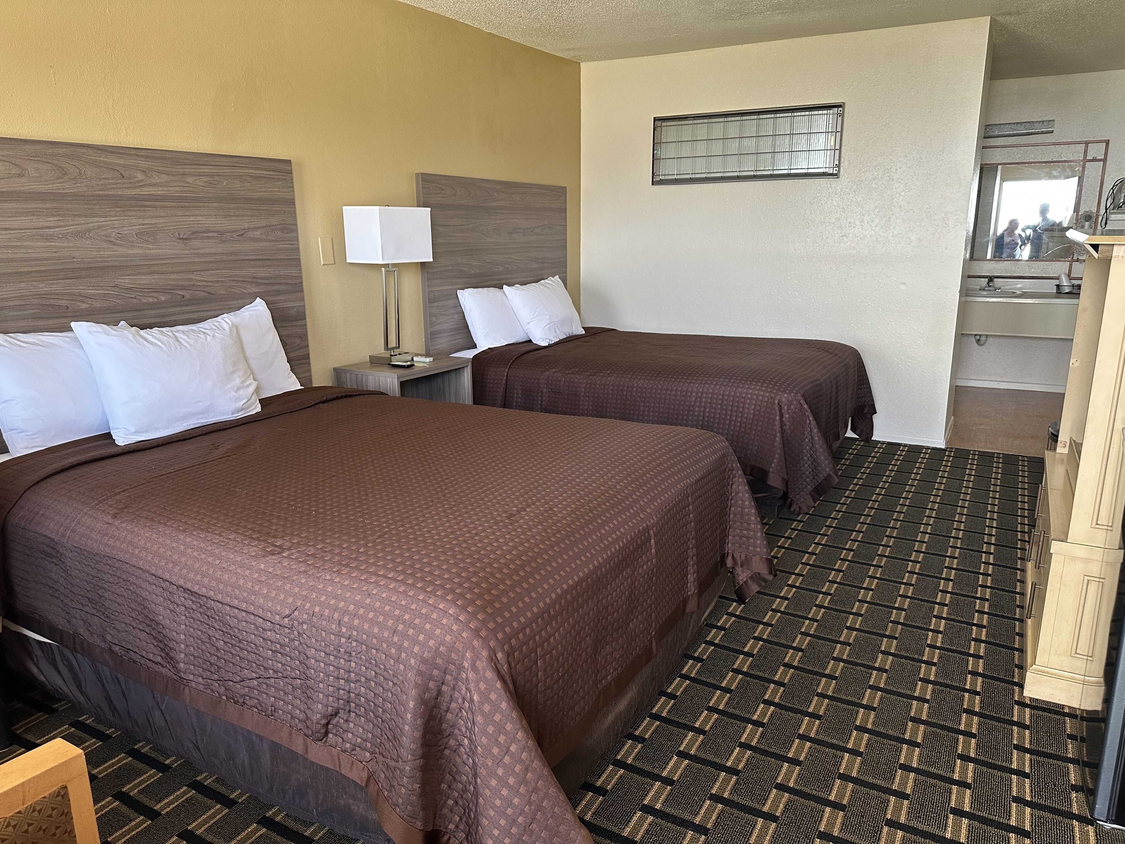 A hotel room with two beds, a nightstand, a lamp, a mirror, and a bathroom visible through an open door.