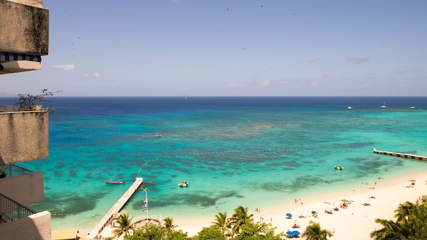 A beach view with clear turquoise water, a pier, and a sandy shore with umbrellas. - Skymiles Beach Suite At  Montego Bay Club Resort (Montego Bay)