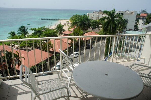 A balcony with a table and chairs, overlooking a beach with palm trees and a pier.