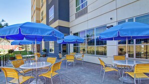 An outdoor patio with round tables and yellow chairs, blue umbrellas, and a building with large windows in the background. - Fairfield Inn and Suites by Marriott Kamloops (Kamloops)