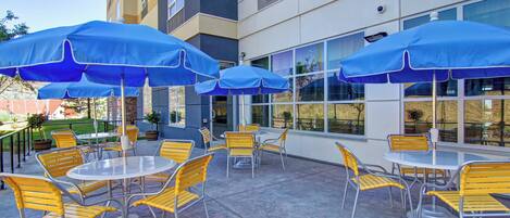 An outdoor patio with round tables and yellow chairs, blue umbrellas, and a building with large windows in the background.