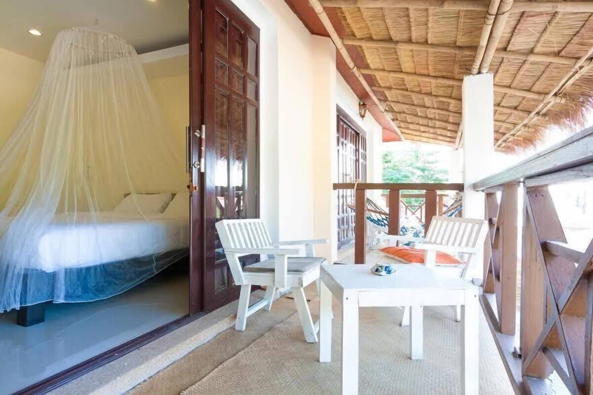 A room with a bed under a mosquito net, a wooden door, and a balcony with white chairs and a table.