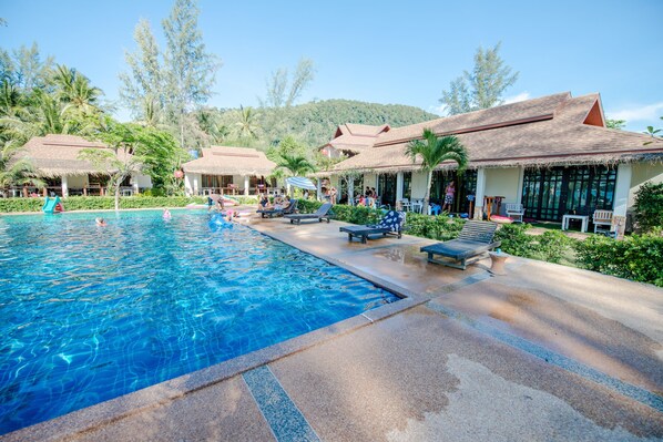 A swimming pool with lounge chairs and umbrellas, surrounded by thatched-roof buildings and lush greenery.