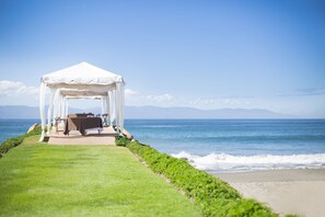 Una carpa blanca con una mesa y sillas instaladas sobre un sendero césped que lleva a la playa.