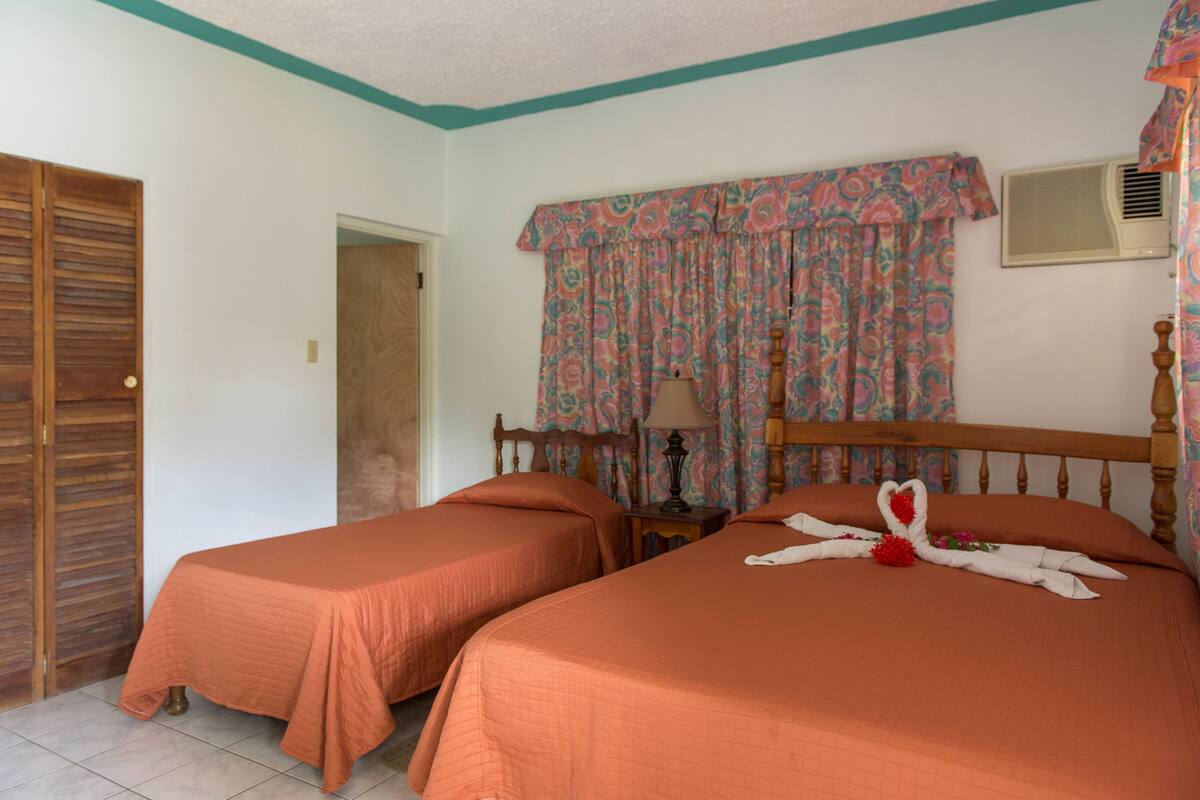A room with two beds, a wooden wardrobe, a door, a window with patterned curtains, and an air conditioning unit.
