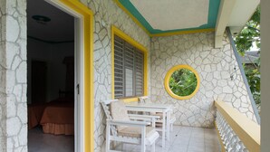 Una habitación con pared de piedra, ventana con contraventanas amarillas, ventanal redondo con vistas verdes y cama con colcha marrón.
