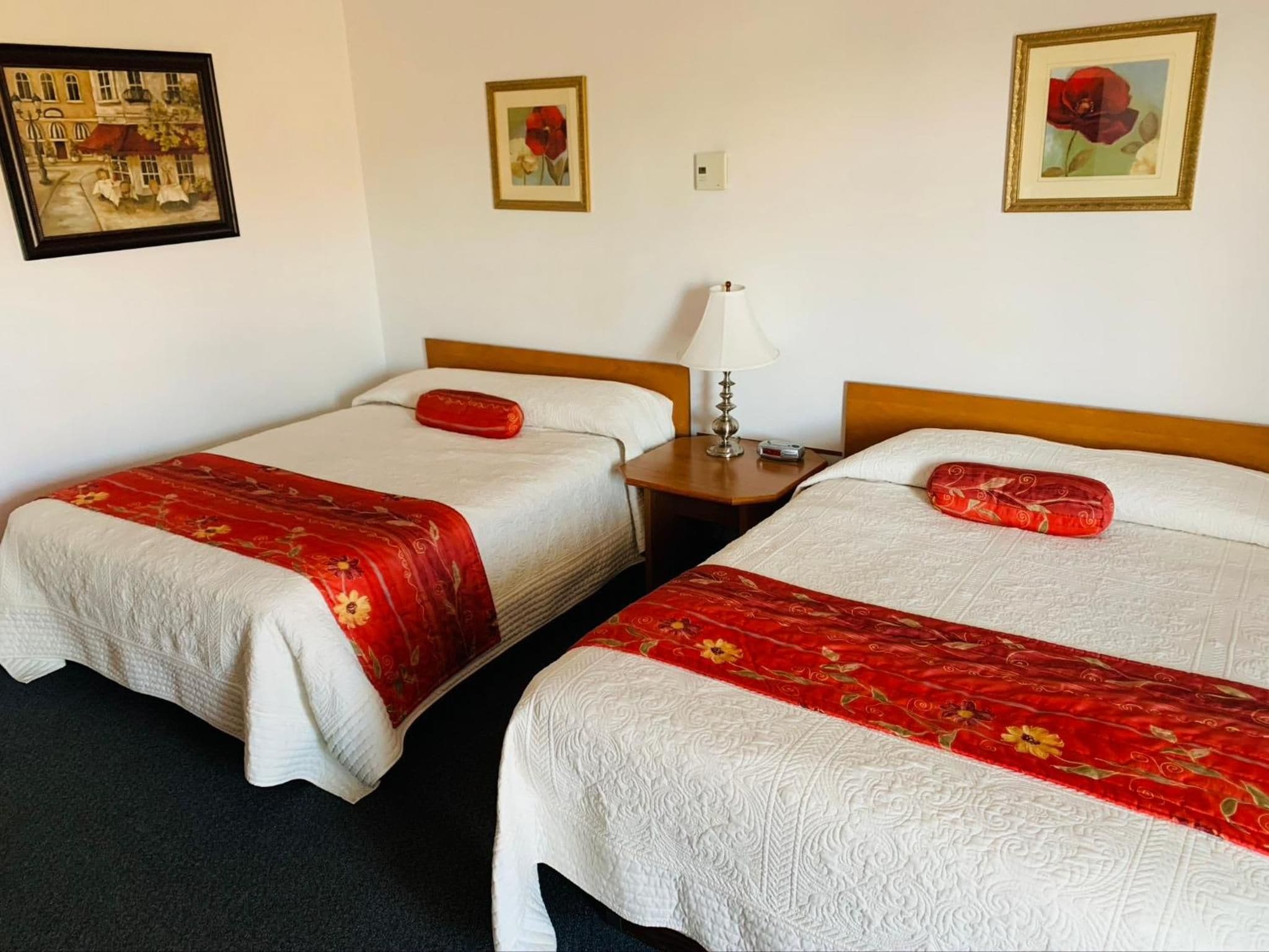 A hotel room with two beds, each with a red floral bedspread and a white pillow. There are two paintings on the walls, one depicting a street scene and the other a red flower.