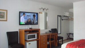 Habitación de hotel con televisión, escritorio, silla, cama, armario y refrigerador pequeño.