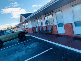 Un motel con un coche verde estacionado al frente, fachada de ladrillo y una fila de sillas afuera.
