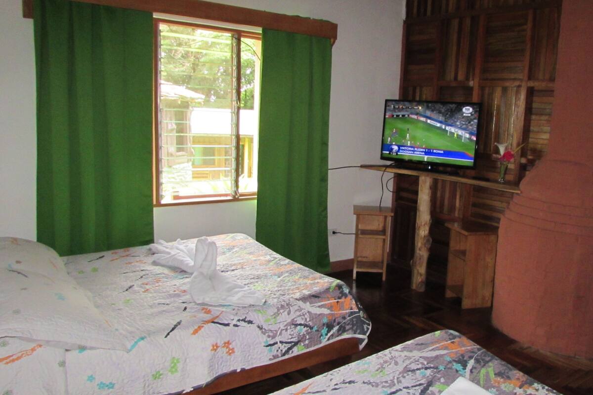 A hotel room with two beds, a television on a wooden stand, a window with green curtains, and a view of the outside.