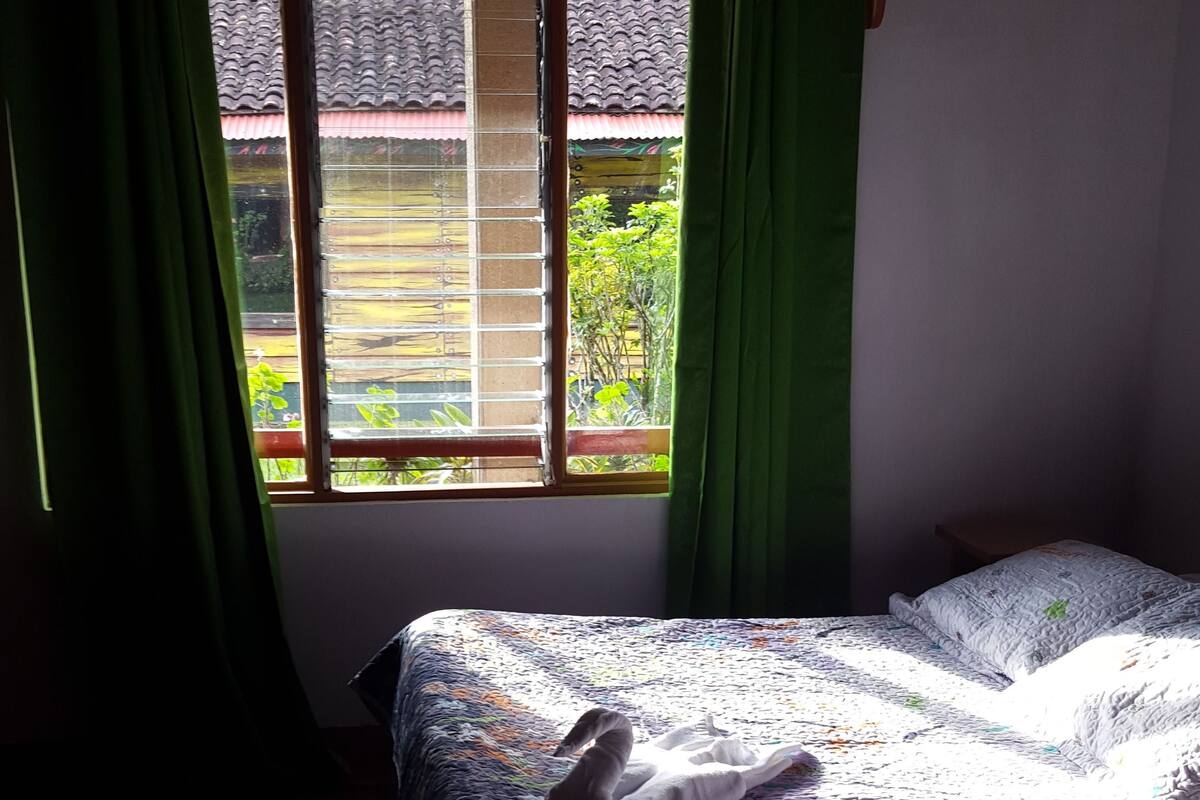 A bedroom with two beds, a window with curtains, and a view of a house and greenery outside.