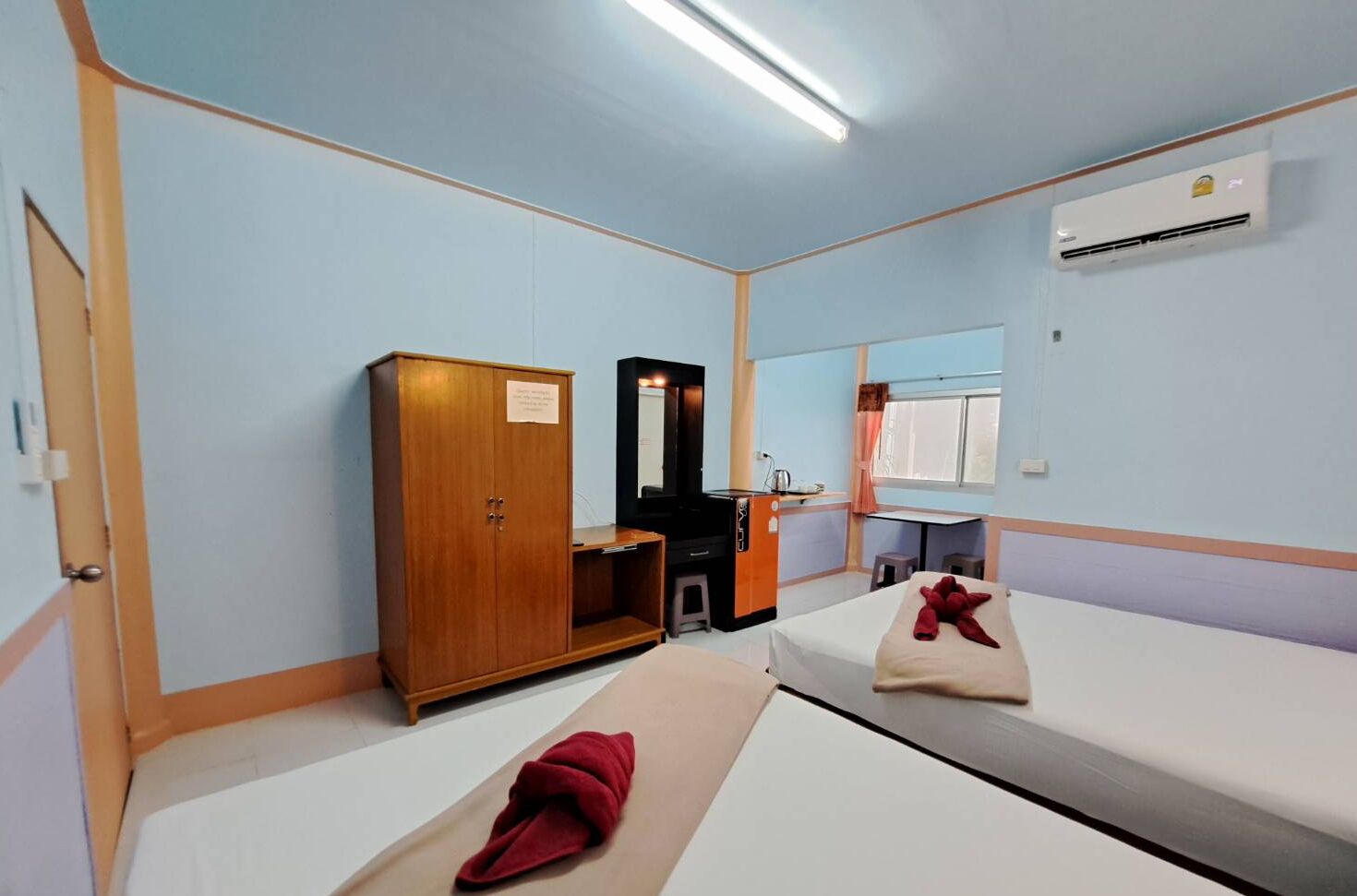 Une petite pièce bien éclairée, avec deux lits, une armoire en bois, un miroir, un bureau avec une chaise et un appareil de climatisation.