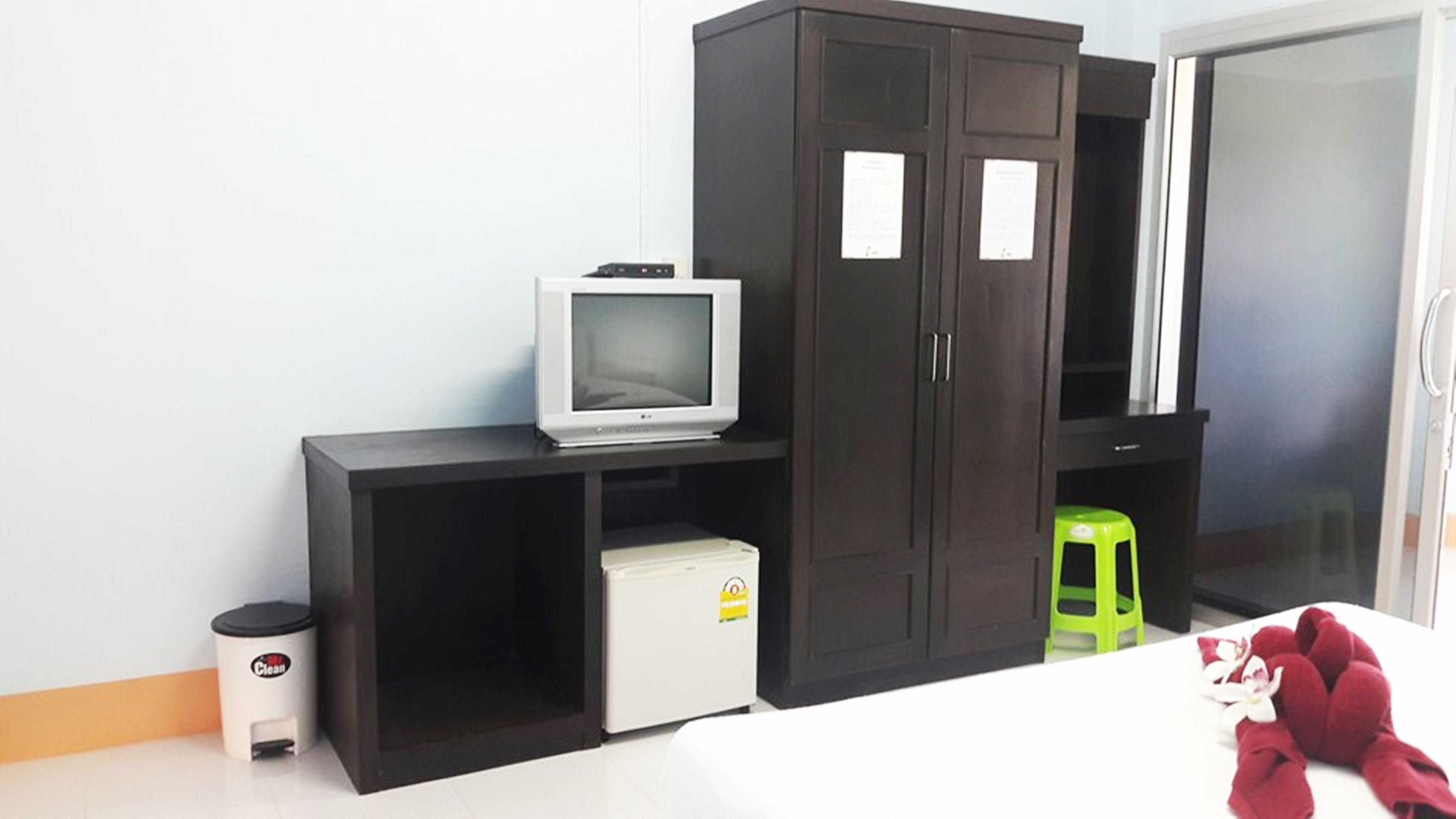 A room with a dark wooden wardrobe, a small white refrigerator, a television on a shelf, a green stool, and a red plush toy on the floor.