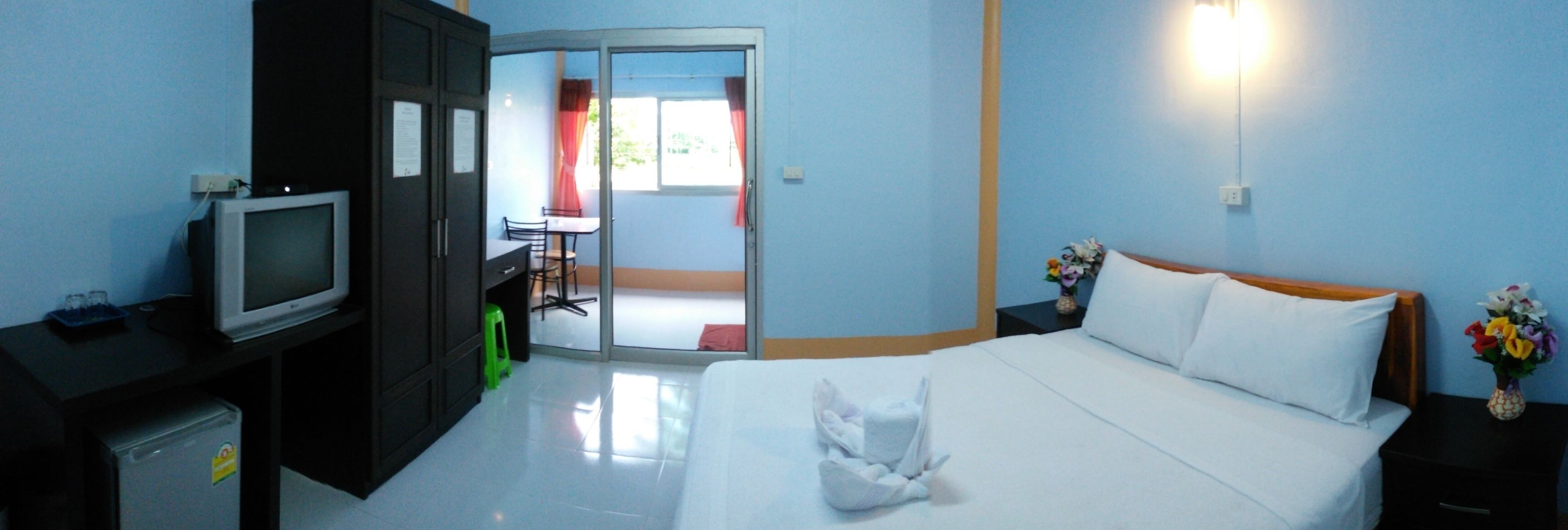 Quarto de hotel com cama, escrivaninha com televisão, pequeno frigorífico, um banquinho verde e porta de correr para uma varanda.