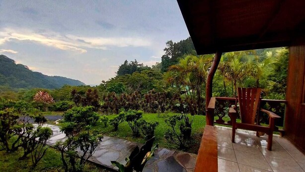 Terraza de madera con una silla, con vista a un jardín exuberante, un río y montañas al fondo.