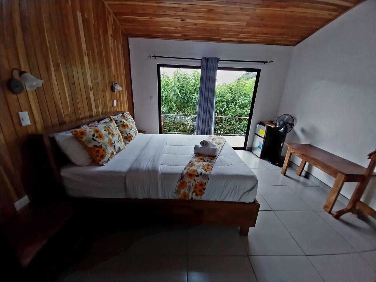 A bedroom with a wooden ceiling, a large bed with floral bedding, a bench, and a view of greenery through a window.