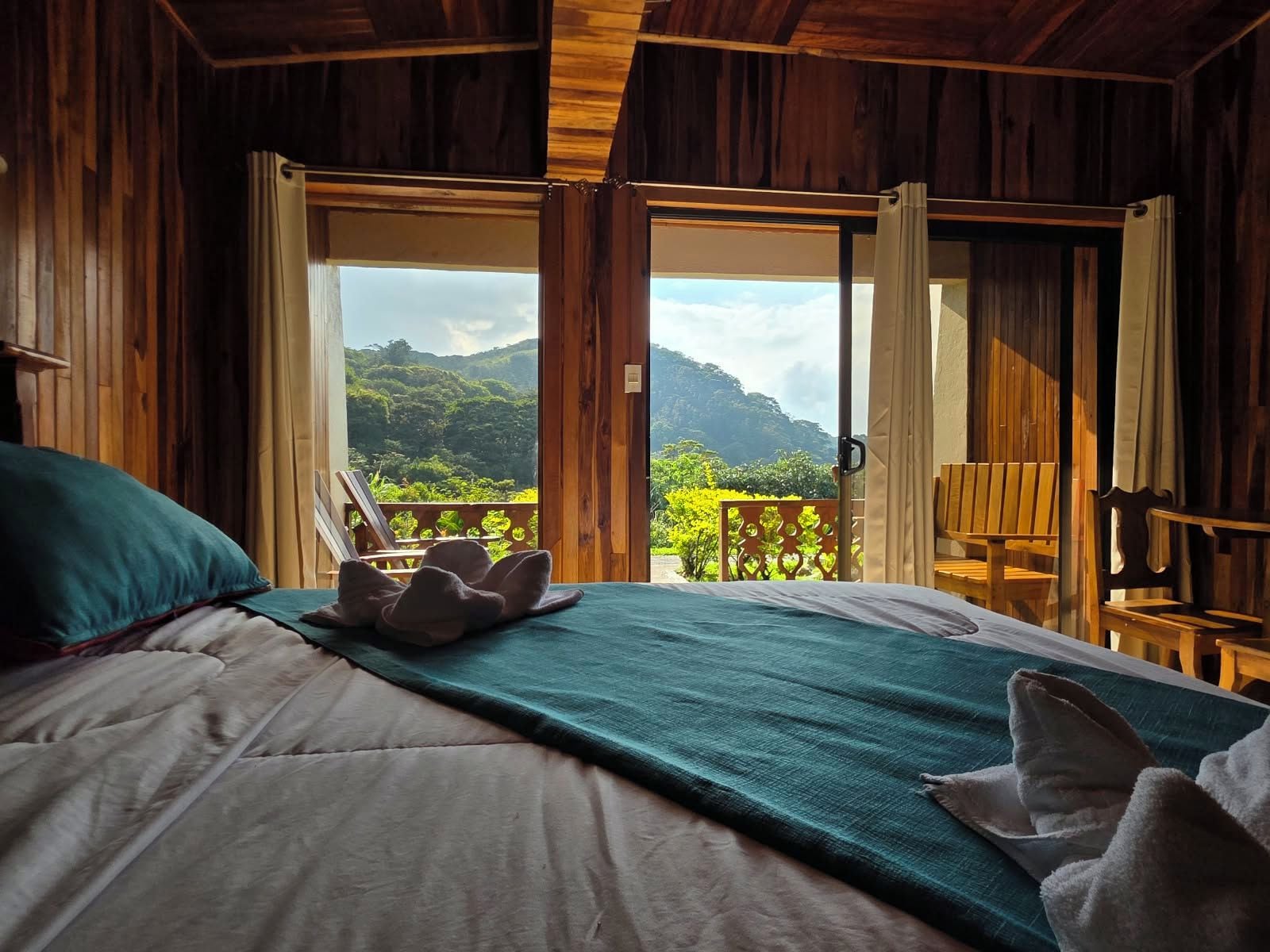 A room with a bed, a view of lush greenery and mountains through open doors, and wooden furniture.