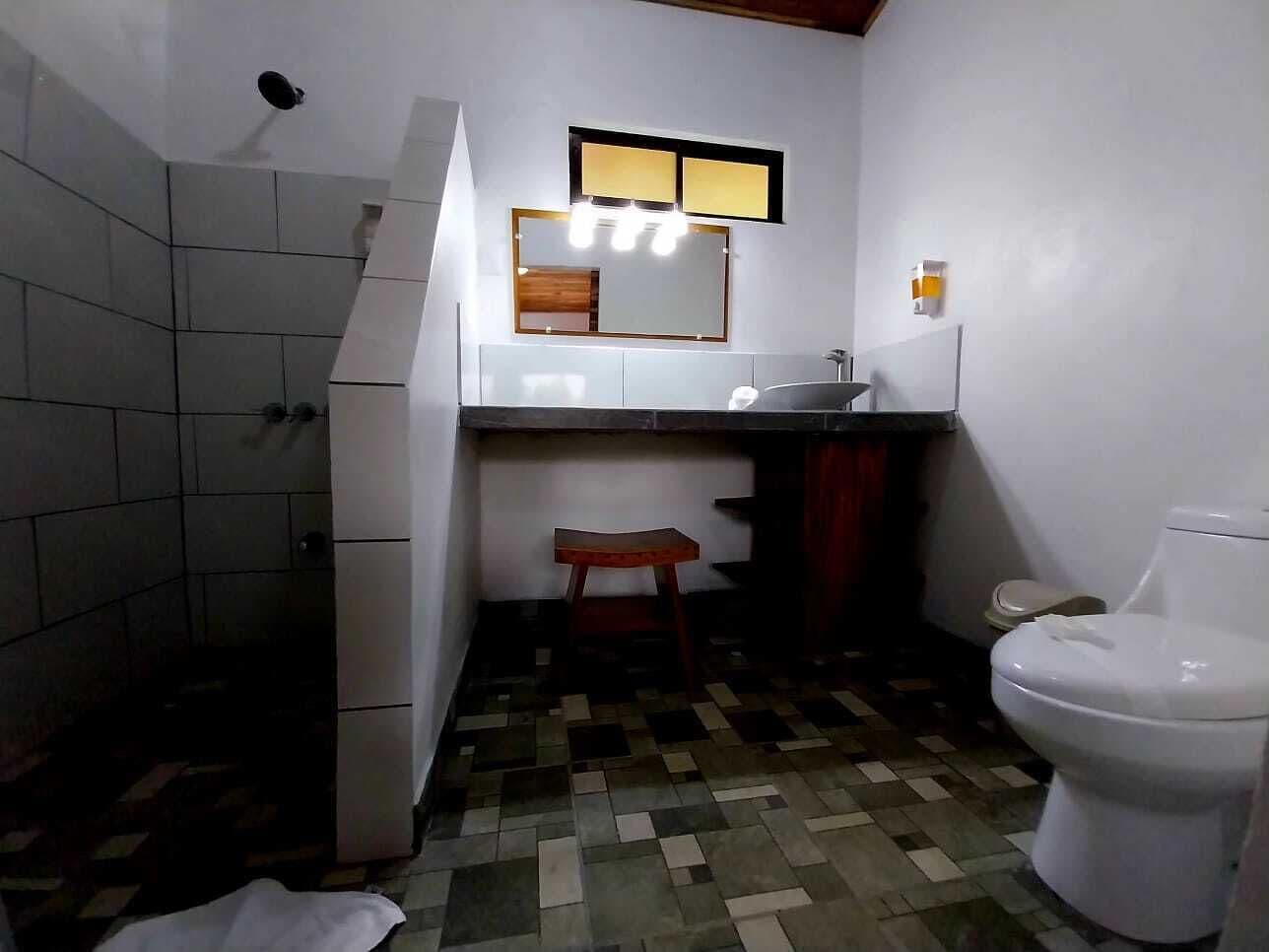 Casa de banho pequena com sanita, lavatório e espelho. Zona de duche à esquerda e chão em mosaico.