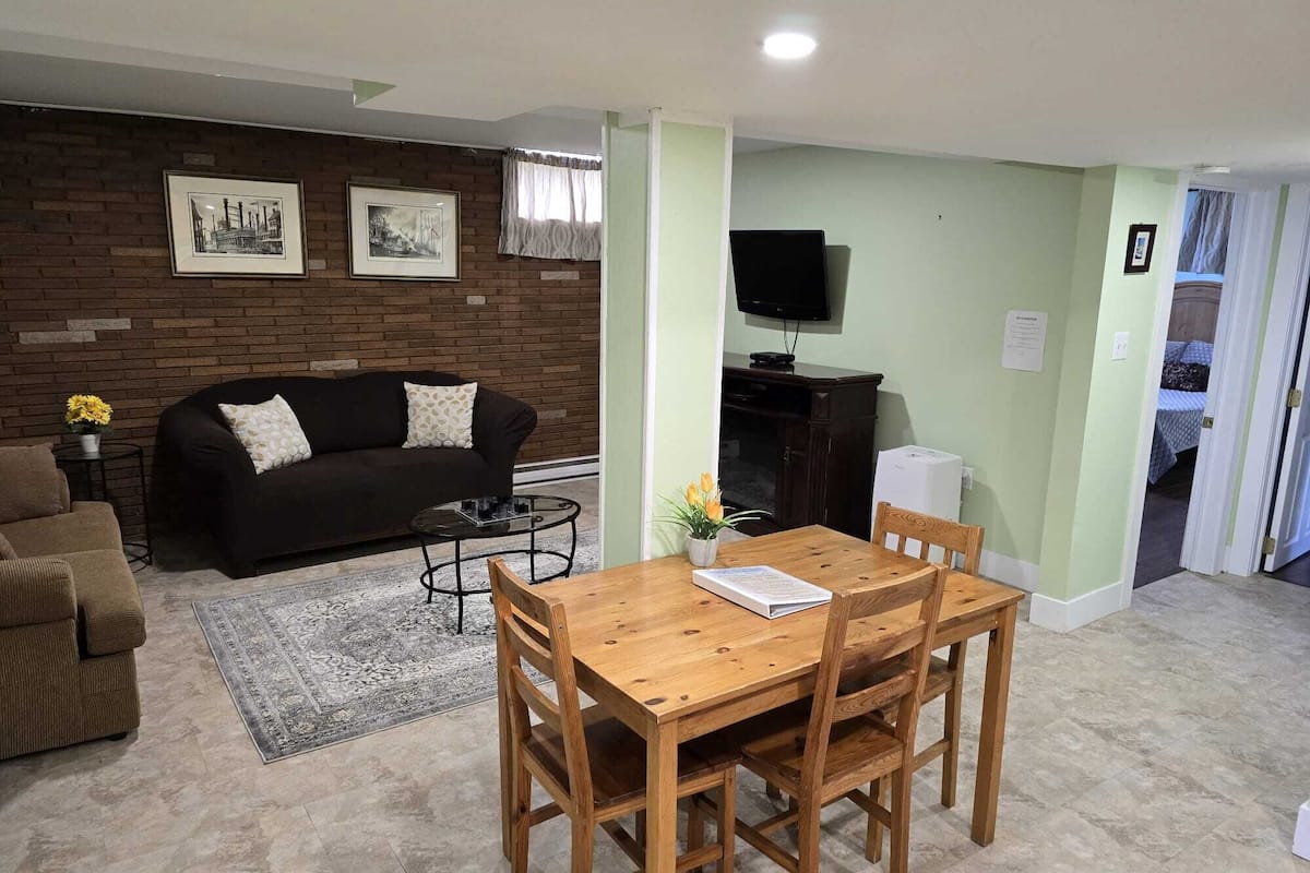 A living room with a dark brown sofa, a wooden dining table with four chairs, a small round coffee table, and a rug. There is a brick wall with framed pictures, a flat-screen TV mounted on the wall, and a door leading to another room.