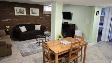 Ein Wohnzimmer mit einem dunkelbraunen Sofa, einem Holzesstisch mit vier Stühlen, einem kleinen runden Couchtisch und einem Teppich. An der Ziegelwand hängen gerahmte Bilder, ein Flachbildfernseher ist an der Wand montiert und eine Tür führt in einen weiteren Raum.