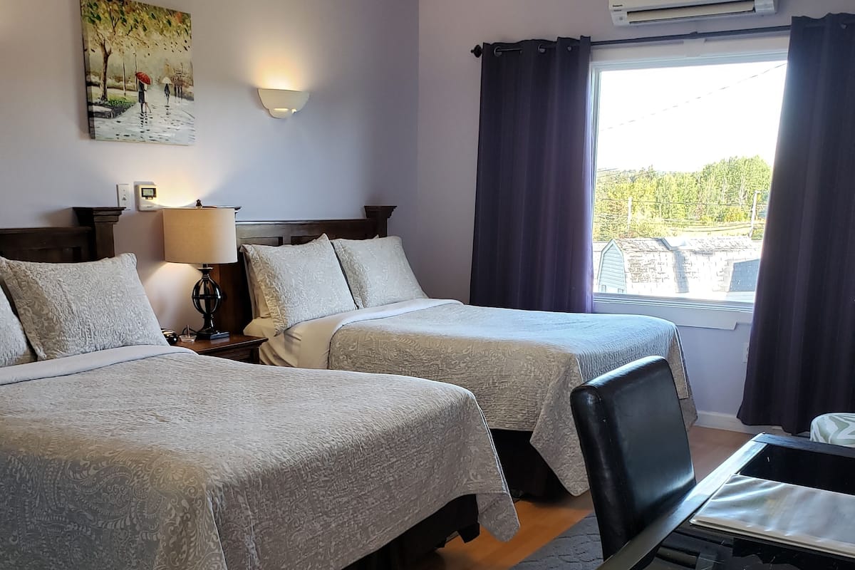A hotel room with two beds, a desk, a lamp, a painting on the wall, and a window with curtains.