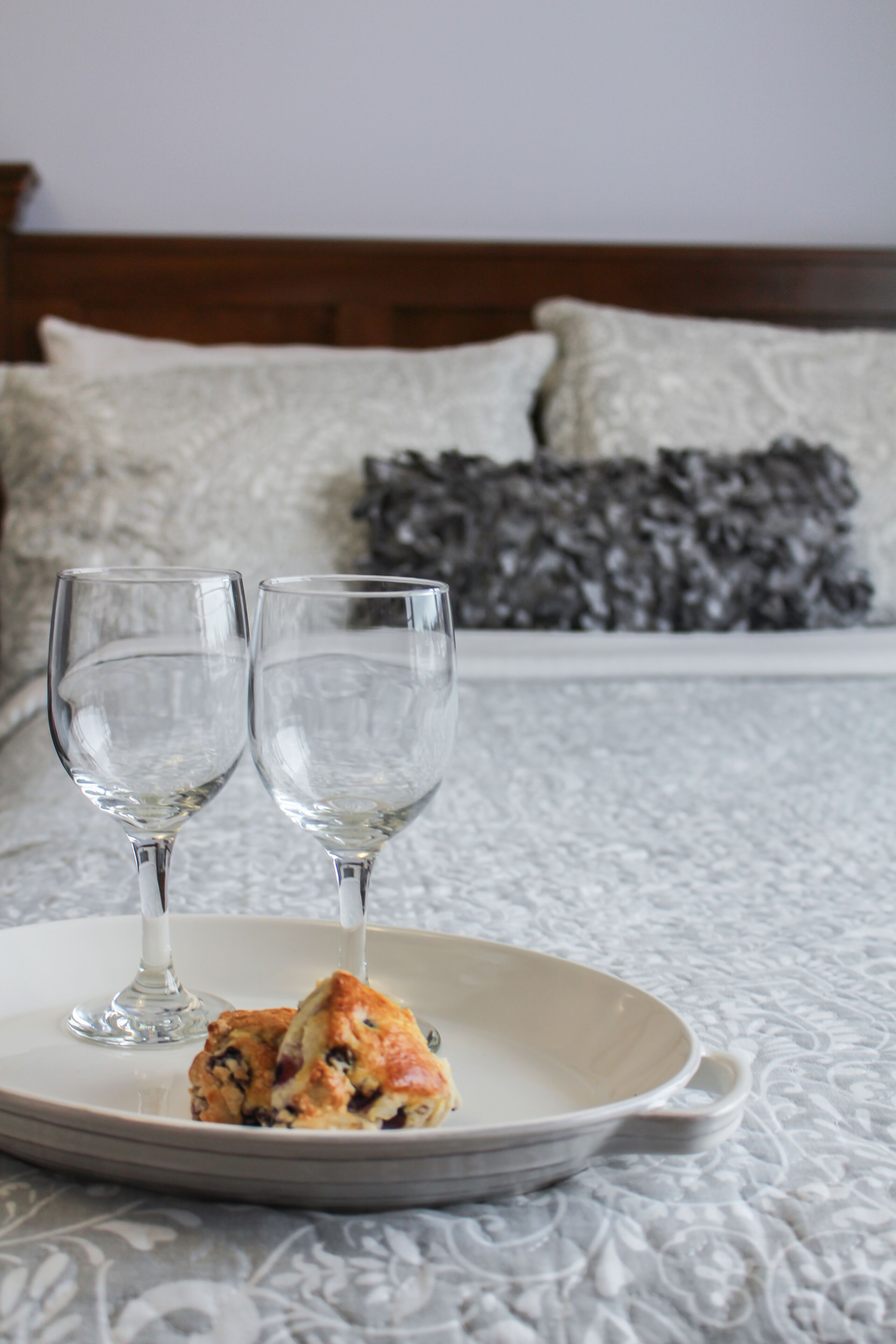 Un letto con una coperta a fascia bianca e grigia, due bicchieri da vino vuoti e un piatto con un po' di cibo su un tavolo.