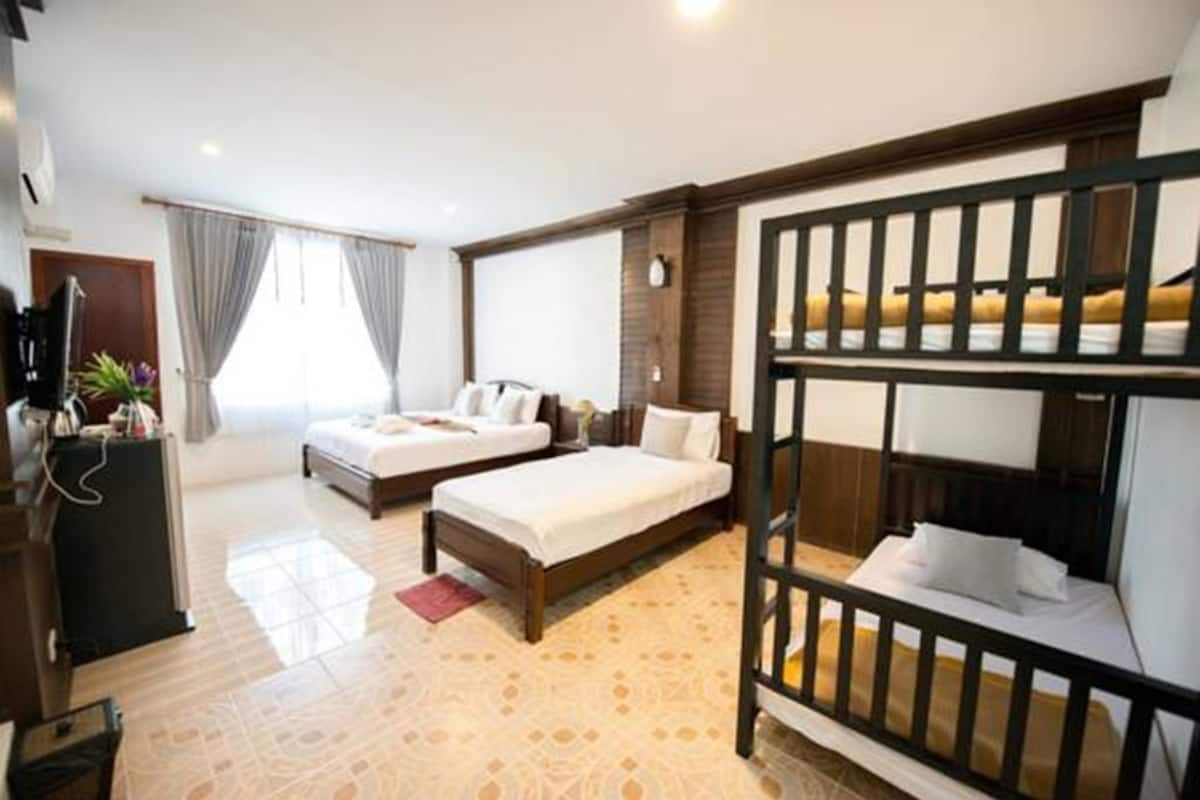 Quarto de hotel com uma cama beliche, uma cama de casal, uma escrivaninha com cadeira e uma grande janela com cortinas.