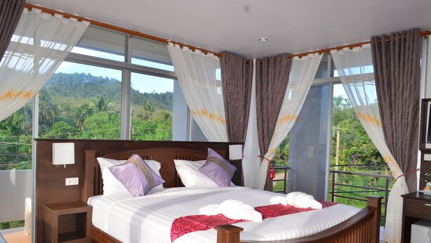 A large bed with a wooden frame, white bedding, and purple pillows. There is a small bedside table with a lamp. The room has large windows with curtains and a view of greenery outside. - Lanta Nice Beach House (Ko Lanta)