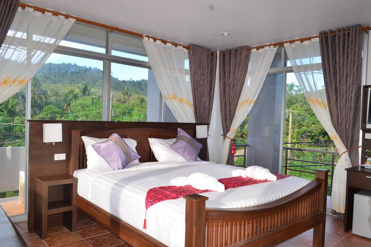 Uma cama grande com estrutura de madeira, roupa de cama branca e almofadas roxas. Há uma pequena mesa de cabeceira com uma luminária. O quarto possui amplas janelas com cortinas e vista para o verde lá fora.