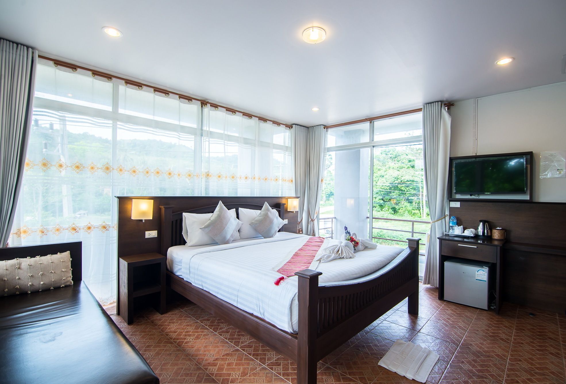 A hotel room with a large bed, a TV, a desk, and a view of greenery outside.