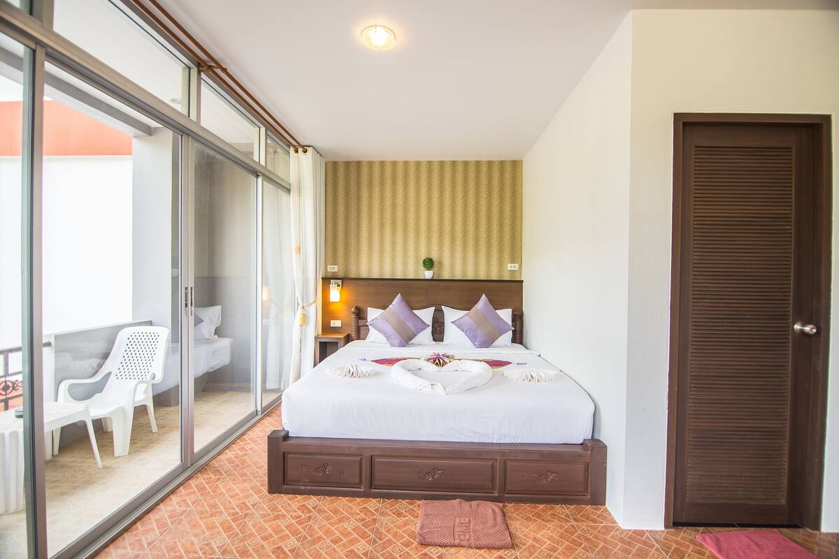 Quarto de hotel com uma cama, uma porta de vidro deslizante que dá para uma varanda, uma porta de madeira e uma pequena mesa de cabeceira com uma luminária.
