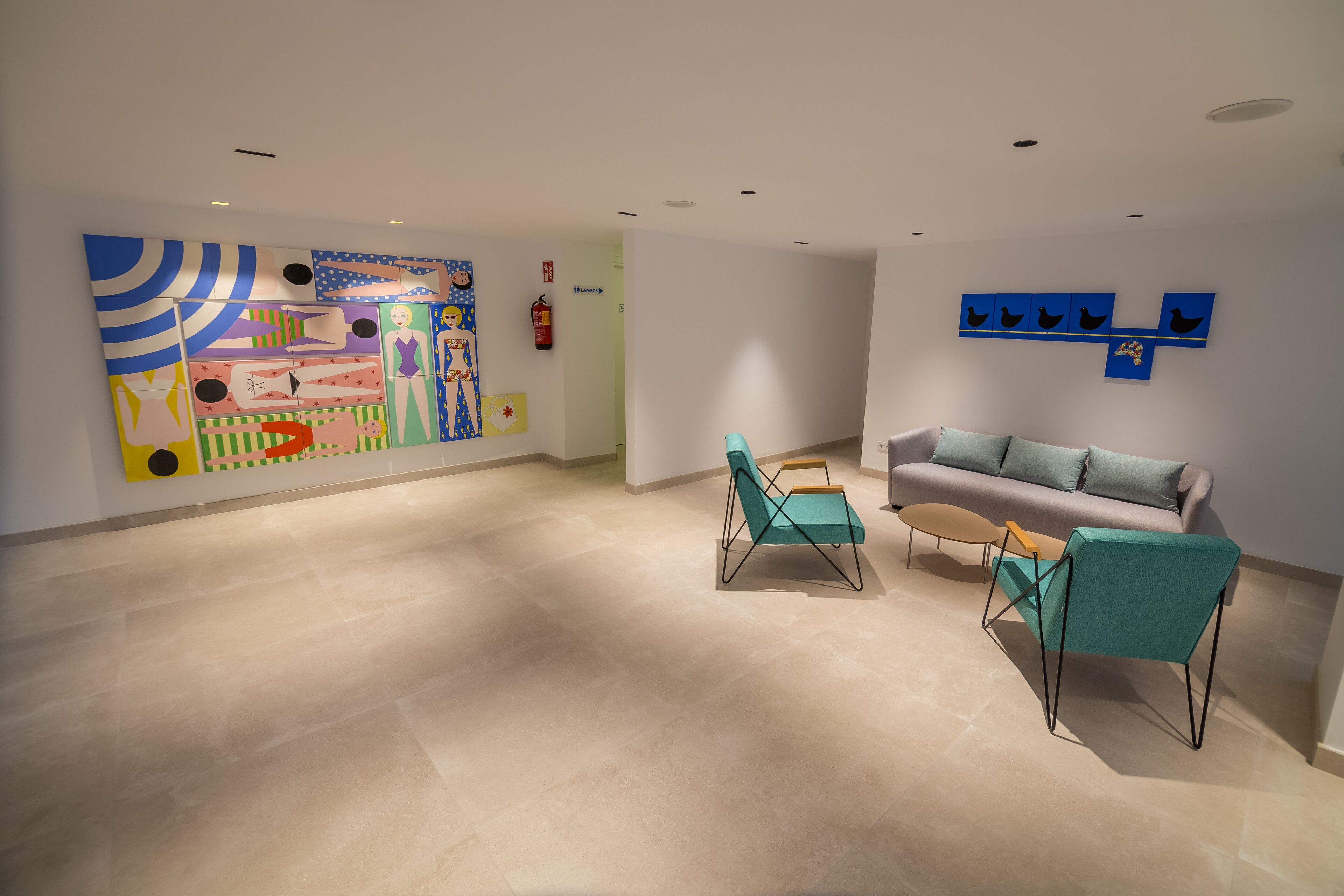 Una habitación amplia con un cuadro abstracto grande, un sofá gris, sillas color turquesa y una pequeña mesa redonda.