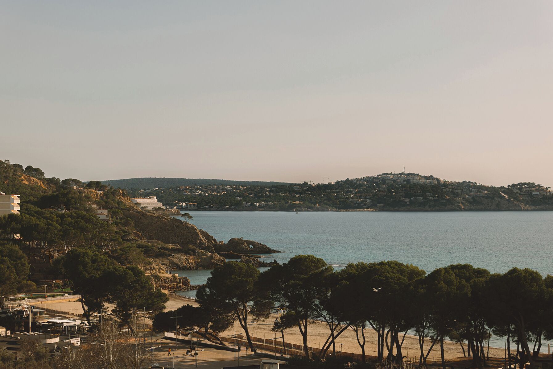 A coastal view with a beach, trees, and a hill with buildings in the background.