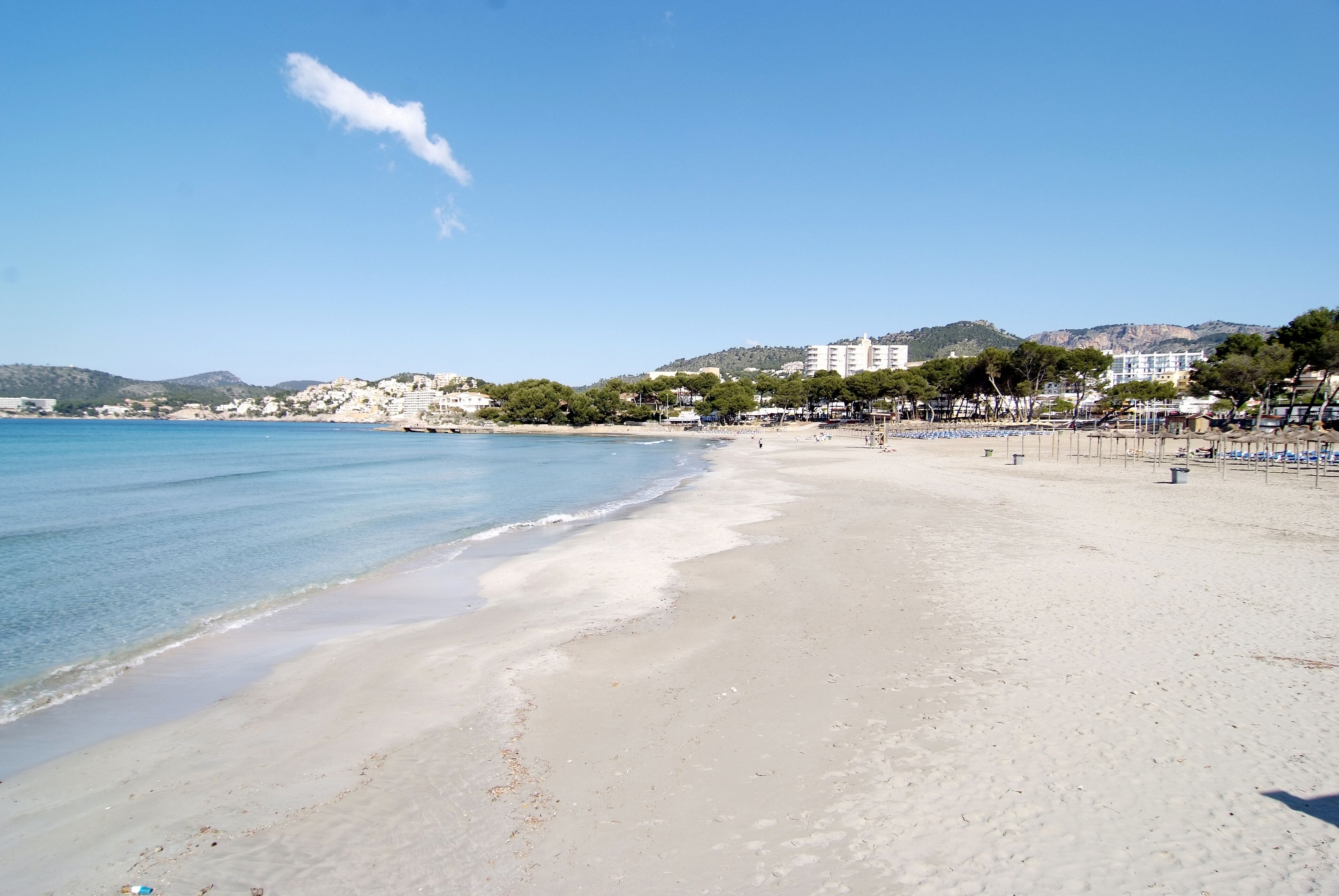 A beach with clear blue water, sandy shore, and buildings in the background.